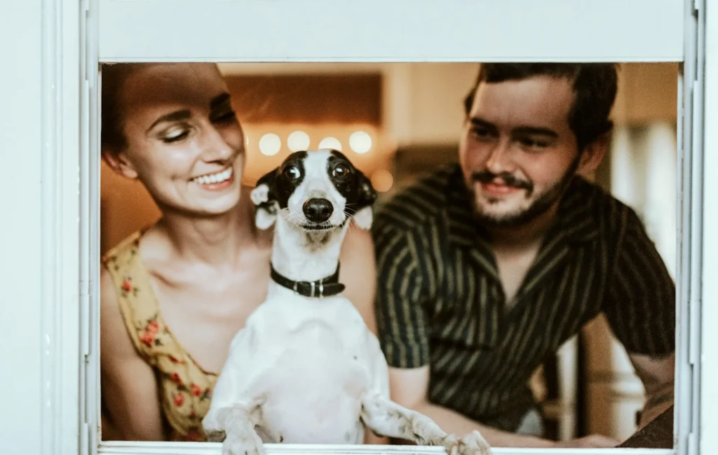 A black and white dog stands between a man and woman, looking through a window frame.